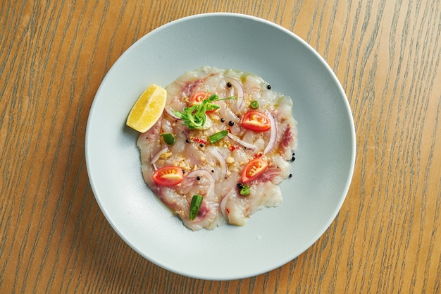 Gekoelde carpaccio van zeebaars met rode ui, cherrytomaatjes en hete chilipepers in een beige kom op een houten muur. Close-up, selectieve aandacht