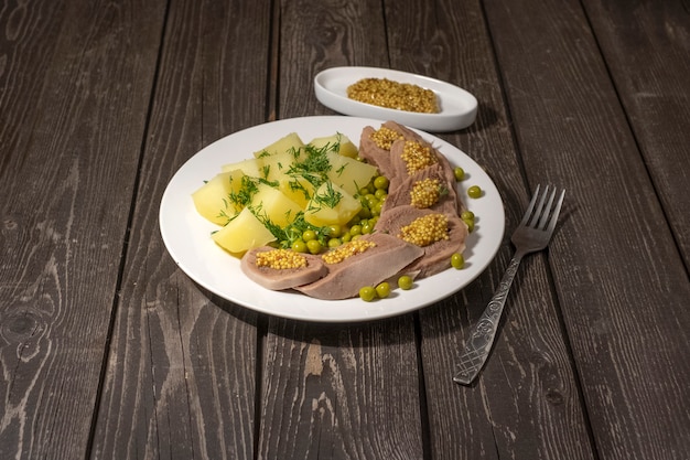 Gekookt rundvlees tong met aardappelen, groene erwten, kruiden en mosterd op donkere houten achtergrond met vork