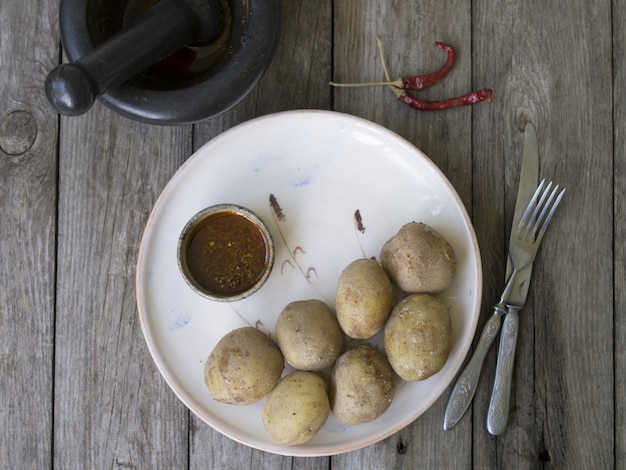 Foto gekookte aardappelen in hun schil. geheel met rode peper, zout en pikante saus op witte plaat, oude houten tafel, rustieke stijl. sluit omhoog, kopieer ruimte. bovenaanzicht.