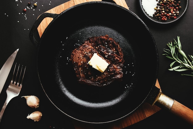 Gekookte ribeye steak op gietijzeren pan en bord met peper, rozemarijn, zout, knoflook, mes, vork, boter op zwarte achtergrond