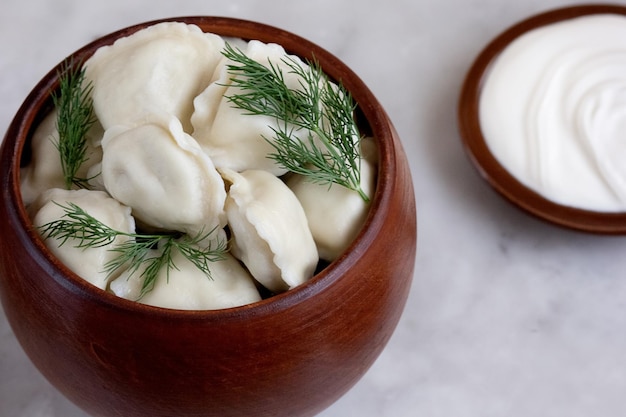 Gekookte Russische pelmeni-vleesknoedels en dille in aarden pot en zure room op marmeren tafel