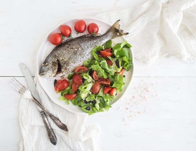 Gekookte zeebrasemvissen met verse groentesalade op ceramische plaat over wit, rustiek houten achtergrondbovenaanzicht