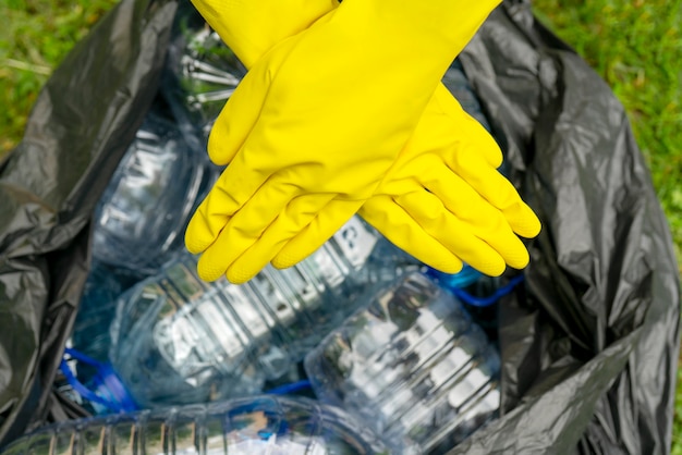 Gekruist handen in gele handschoenen tegen een zwarte pklastic tas met lege plastic flessen. Plastic verbod.