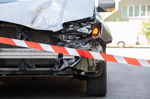 Foto gekruiste rood-witte waarschuwingsband voor vernietigd auto-ongeluk kapotte motorkap van auto op omheinde weg