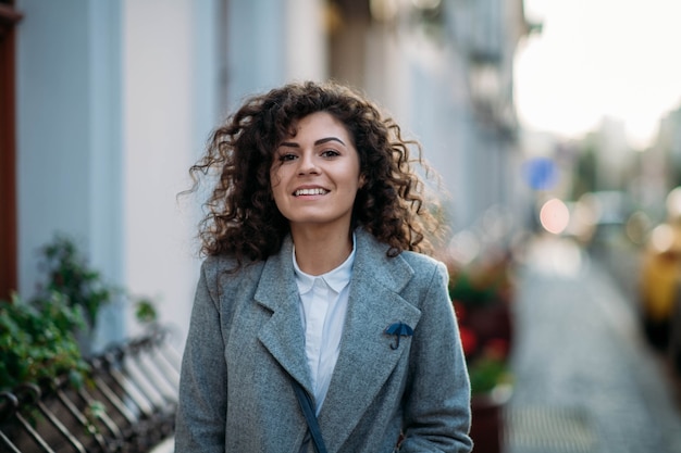 gekrulde jonge vrouw in een grijze jas loopt in de herfst door de stad