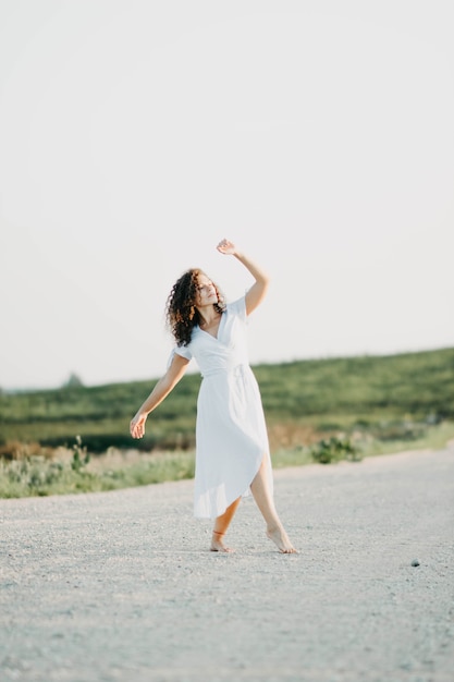 gekrulde jonge vrouw romantisch dansen in een blauwe jurk op een zandweg bij zonsondergang