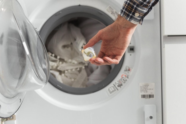 Gel caps in hand for washing machine, liquid coloured detergent and conditioner with full uploaded washer by white clothes in background