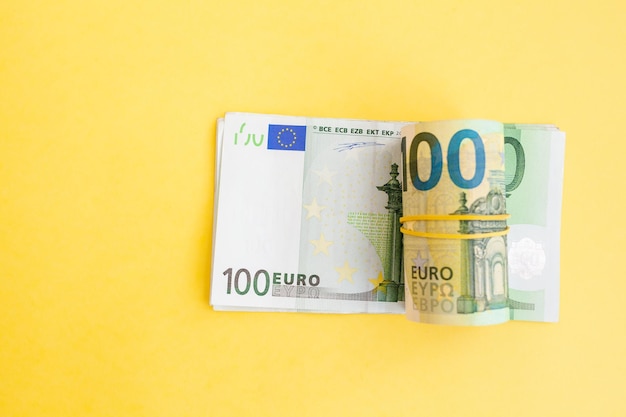 Geldrol van eurobankbiljetten met elastische band op een papieren biljet van 100 euro op een gele achtergrond.