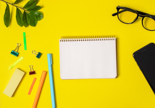 Gele achtergrond, witte notebook voor records, telefoon, computer bril, takje planten, pennen, potloden.