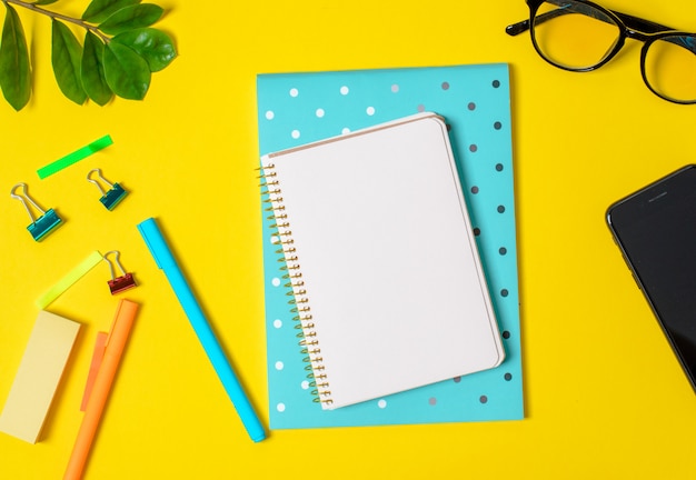 Gele achtergrond, witte notebook voor records, telefoon, computer bril, takje planten, pennen, potloden.