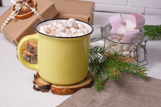 Gele kop met cacao versierd met marshmallows op een standaard gemaakt van gezaagd hout, een pot marshmallows, een sparrentak en kerstcadeaus op de achtergrond