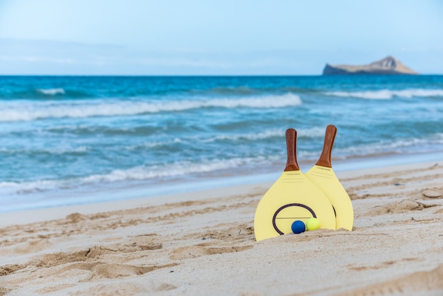 Gele strandpeddel en ballen op een zandig strand in Hawaï met golven en een eiland op de achtergrond