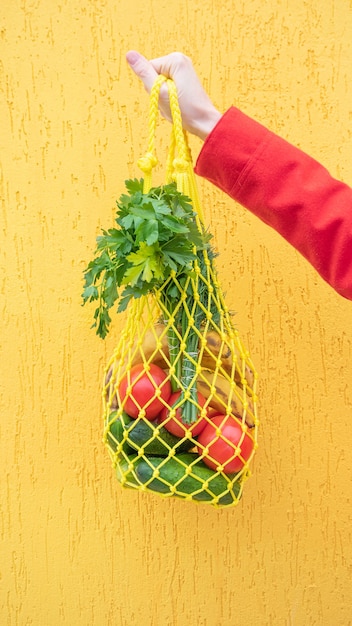 Gele string tas met komkommers, tomaten, bananen en kruiden in de hand close-up. Heldere foto in rode, gele, groene tinten. Duurzaamheid, nul afval, plasticvrij concept, vegetarisme, gezonde voeding.