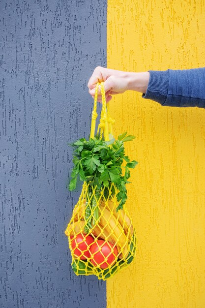 Foto gele string tas met komkommers, tomaten, bananen en kruiden in de hand close-up. heldere foto in rode, gele, groene tinten. duurzaamheid, nul afval, plasticvrij concept, vegetarisme, gezonde voeding.