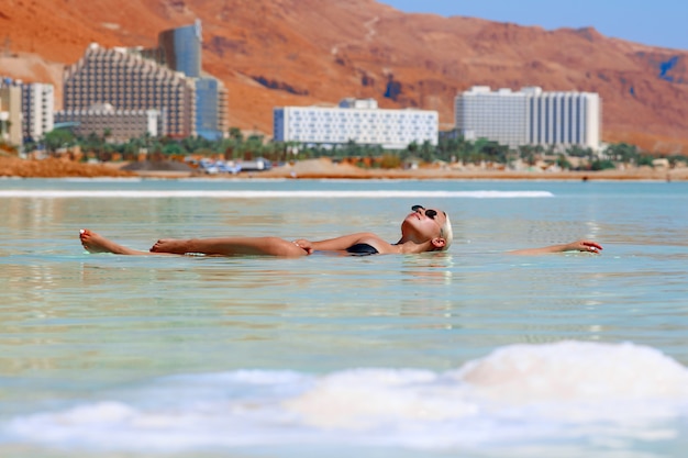 Gelooid sexy blond meisje in een zwarte zwembroek en zonnebril zwemt in het water van de Dode Zee in Israël