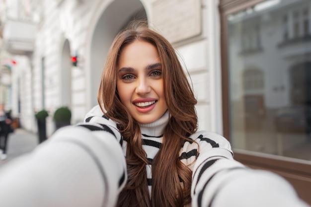 Foto geluk jonge mooie vrouw met schattige glimlach in mode gestreepte trui geniet van reizen en selfie maken op smartphone in de buurt van een wit vintage gebouw in de europese stad