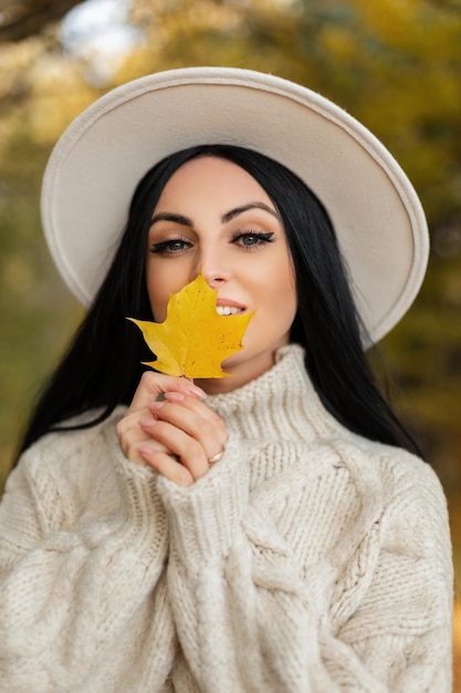 Geluk mooi meisje met glimlach in vintage mode trui en witte ronde hoed houdt een geel herfstblad vast en loopt in het herfstpark Trendy vrouw kijkt naar de camera