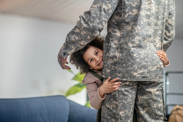 Geluk. Schattig gelukkig klein meisje met donker krullend haar knuffelen vader in militair uniform staande thuis in de kamer