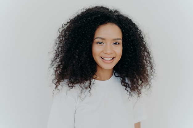 Geluk, welzijn en positieve emoties concept Headshot van aangenaam ogende dame met fris haar glimlacht en lacht heeft een gezonde huid draagt casual t-shirtmodellen tegen witte studiomuur