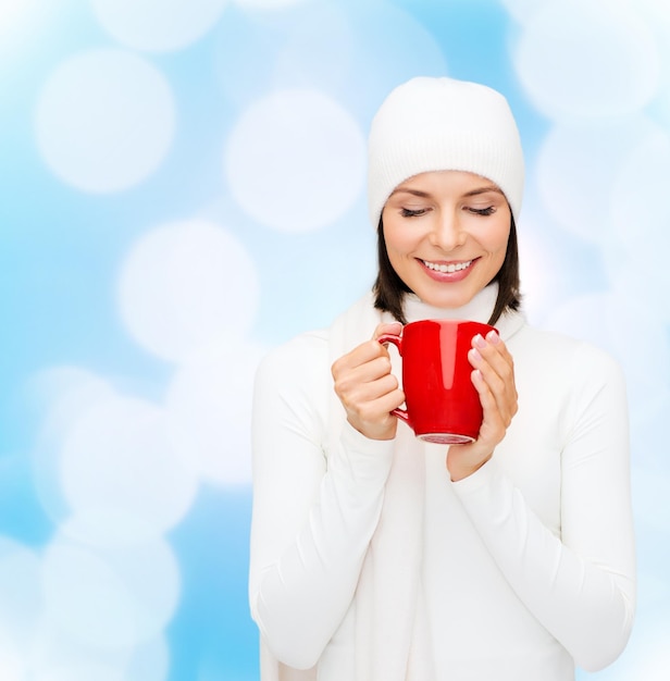 Foto geluk, wintervakantie, kerstmis, dranken en mensenconcept - glimlachende jonge vrouw in witte warme kleren met rode kop over blauwe lichtenachtergrond