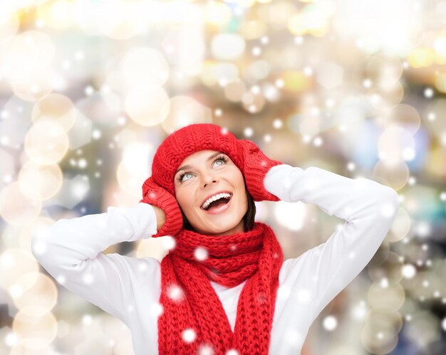 geluk, wintervakantie, kerstmis en mensenconcept - glimlachende jonge vrouw in rode hoed, sjaal en wanten over lichtenachtergrond