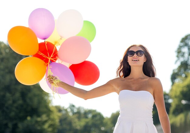 geluk, zomer, vakantie en mensenconcept - glimlachende jonge vrouw die zonnebril met ballons in park draagt