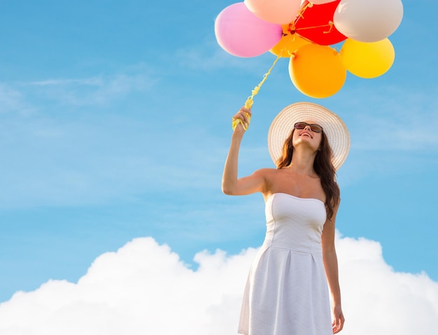 geluk, zomer, vakantie en mensenconcept - glimlachende jonge vrouw die zonnebril met ballons over blauwe hemel en wolkenachtergrond draagt