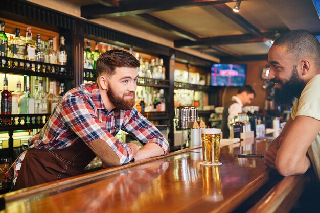 Gelukkig aantrekkelijke jonge barman glas bier geven en praten met jonge man