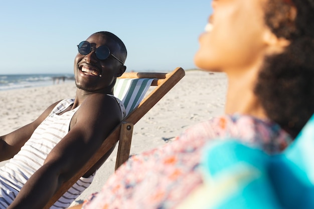 Gelukkig Afrikaans Amerikaans echtpaar in zonnebril zittend in ligstoelen lachend op zonnig strand. Zomer, ontspanning en vakantie, onveranderd.