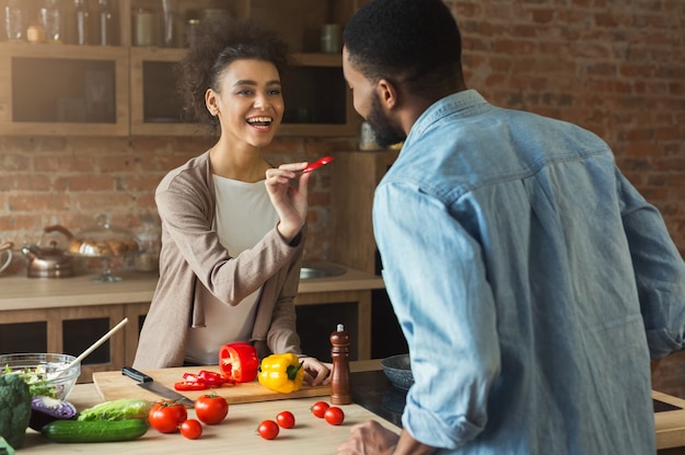 Gelukkig Afrikaans-Amerikaans stel dat thuis gezond voedsel kookt en proeft in hun loftkeuken. Vrouw en man die wijn drinken. Groentesalade bereiden.