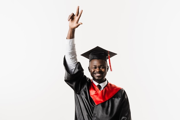 Gelukkig Afrikaanse student in zwarte afstudeerjurk en pet steekt zijn handen boven het hoofd op witte achtergrond Afgestudeerde Afrikaanse man studeert af en viert academische prestatie