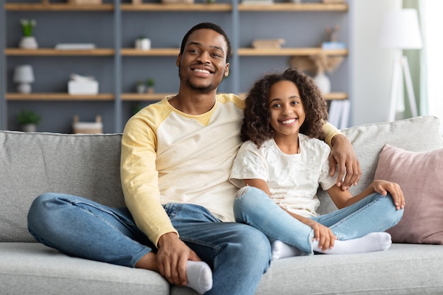 Gelukkig Afro-Amerikaanse familie schoolmeisje jongen en vader tijd samen doorbrengen thuis, vrolijke zwarte vader en dochter zittend op de bank en omarmen, glimlachend in de camera, kopieer ruimte