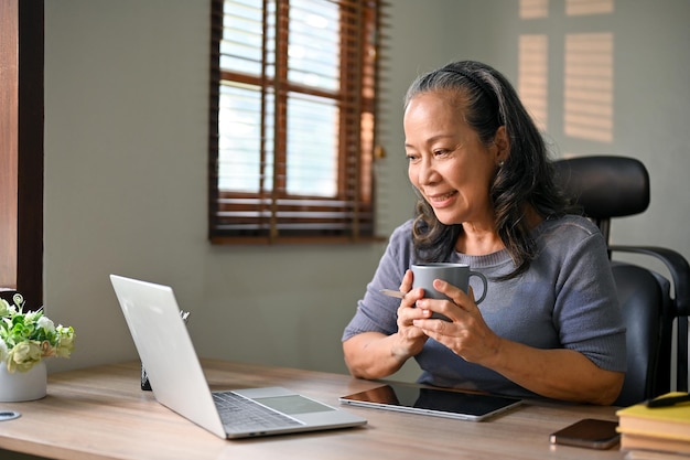 Gelukkig Asianage zakenvrouw nippen aan koffie tijdens het werken aan haar zakelijke taken op laptop