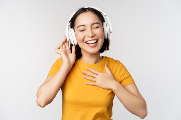 Gelukkig Aziatisch meisje dansen luisteren muziek op koptelefoon en glimlachend staande in gele t-shirt tegen witte achtergrond