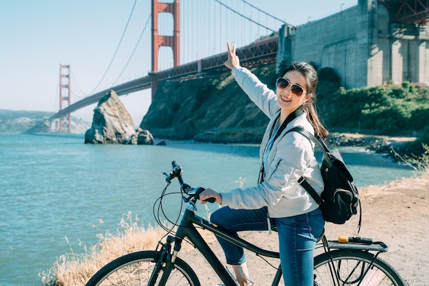 gelukkig aziatisch meisje toerist kijkt naar de camera met een handgebaar van het vredesteken op de fiets met golden gate bridge en blauwe zee op de achtergrond in San Francisco, VS