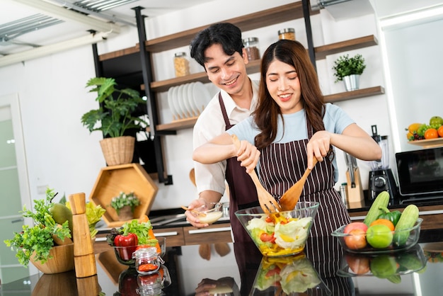 Gelukkig Aziatisch paar dat schorten draagt die de vegetarische salade met houten spatels gooien Het voorbereiden van een gezonde salade met verse groenten zoals worteltomaatkool en groene eik in de huiskeuken