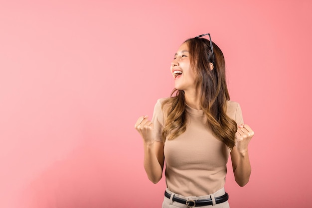 Gelukkig Aziatisch portret mooie schattige jonge vrouw draagt een bril maakt opgeheven hand omhoog om haar winnende succesgebaar te vieren, studio-opname geïsoleerde roze achtergrond, vrouwelijk opgewonden zeg ja! met kopie ruimte