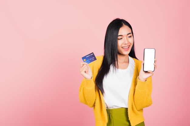 Gelukkig Aziatisch portret mooie schattige jonge vrouw opgewonden glimlachend houd mobiele telefoon en plastic debet creditcard bankkaart, studio opname geïsoleerd op roze achtergrond, vrouw met behulp van smartphone online winkelen