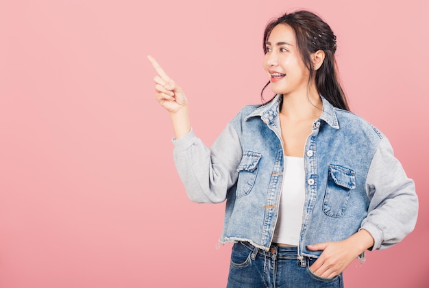 Gelukkig Aziatisch portret mooie schattige jonge vrouw staande glimlachend wijzen vinger lege ruimte op zoek naar camera, studio opname geïsoleerd op roze achtergrond, Thaise vrouwelijke wijzende index uit met kopie ruimte