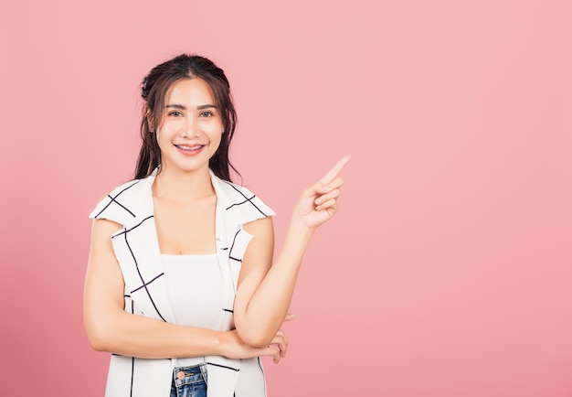 Gelukkig Aziatisch portret mooie schattige jonge vrouw staande glimlachend wijzen vinger lege ruimte op zoek naar camera, studio opname geïsoleerd op roze achtergrond, Thaise vrouwelijke wijzende index uit met kopie ruimte