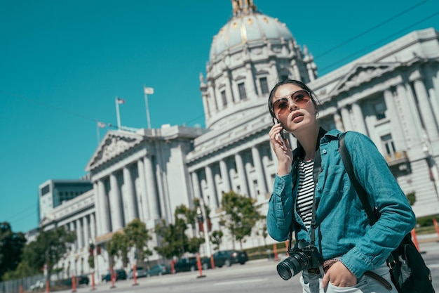 Gelukkig Aziatische jonge vrouw in zonnebril bellen op smartphone. beroemde stadhuis van san francisco op de achtergrond onder de blauwe hemel overdag. elegante toeristische meisje met camera praten op mobiele telefoon.