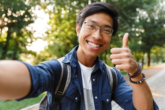 Gelukkig Aziatische mannelijke student in brillen selfie maken en duim opdagen tijdens het kijken naar de camera buitenshuis