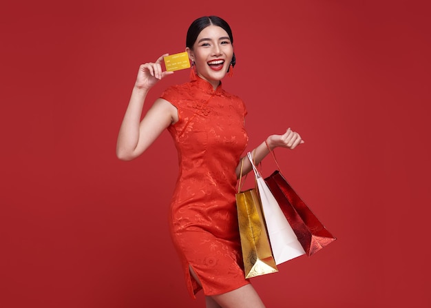 Gelukkig Aziatische shopaholic vrouw dragen traditionele cheongsam qipao jurk met creditcard en boodschappentas geïsoleerd op rode achtergrond. Gelukkig Chinees nieuwjaar