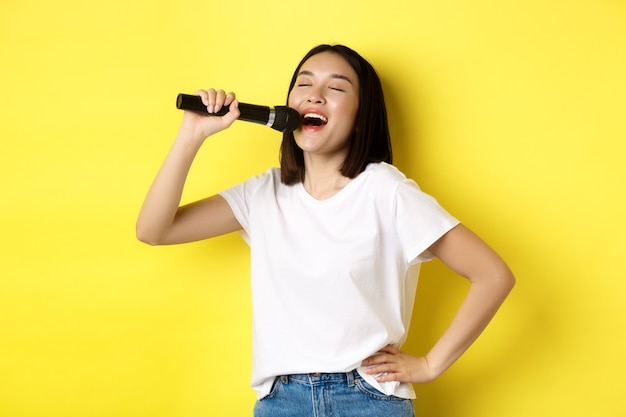 Gelukkig Aziatische vrouw zingen lied in karaoke, microfoon houden, staande over geel.