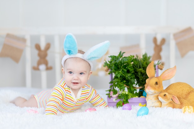 Gelukkig babyjongen met konijnenoren op zijn hoofd liggend met een konijn op het bed met paaseieren, leuke grappige glimlachende kleine baby.