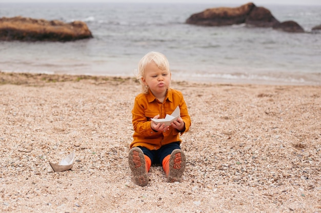 Gelukkig babyjongen spelen met papieren boot op het strand in de herfsttijd