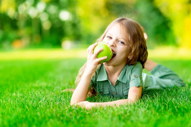 Gelukkig babymeisje in de zomer op het gazon bijt een groene appel met gezonde tanden op het gras en glimlacht, ruimte voor tekst