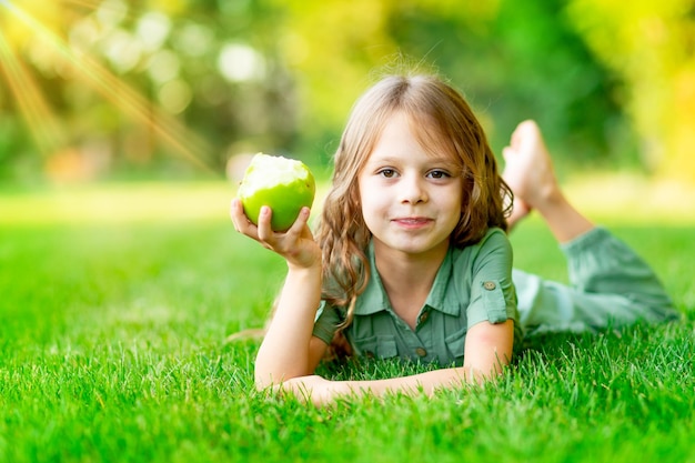 Gelukkig babymeisje in de zomer op het gazon bijt een groene appel met gezonde tanden op het gras en lacht ruimte voor tekst