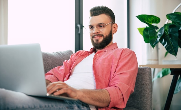 Gelukkig bebaarde aantrekkelijke jonge man in glazen en vrijetijdskleding met een laptop op de bank in huis of appartement is relaxin of werkt