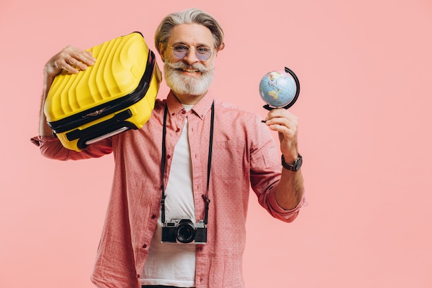 Gelukkig bebaarde man in zonnebril met een camera houdt een koffer en een wereldbol en bereidt zich voor op de reis op roze.